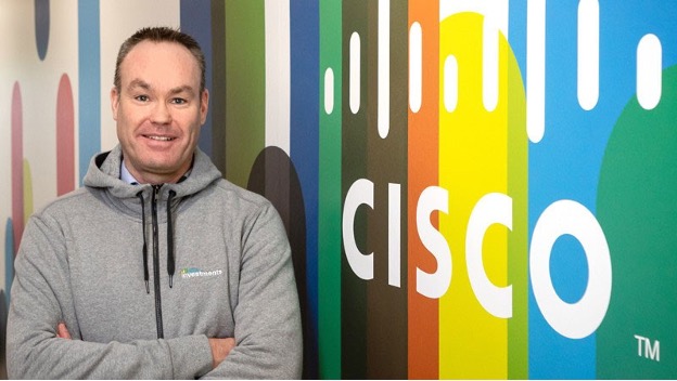 Imagem de Keith ao lado de um mural do logotipo da Cisco