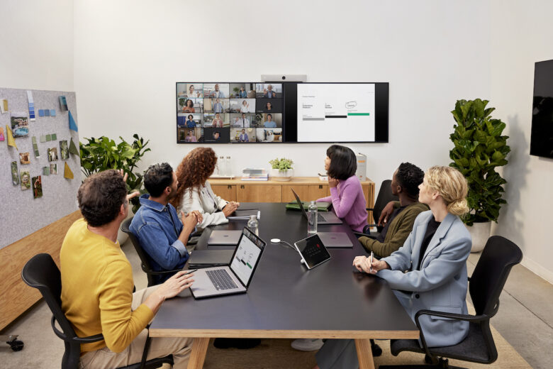 Seis colegas em uma mesa. Eles estão usando tecnologia de videoconferência para interagir com seus colegas a partir de um monitor fixado na parede.