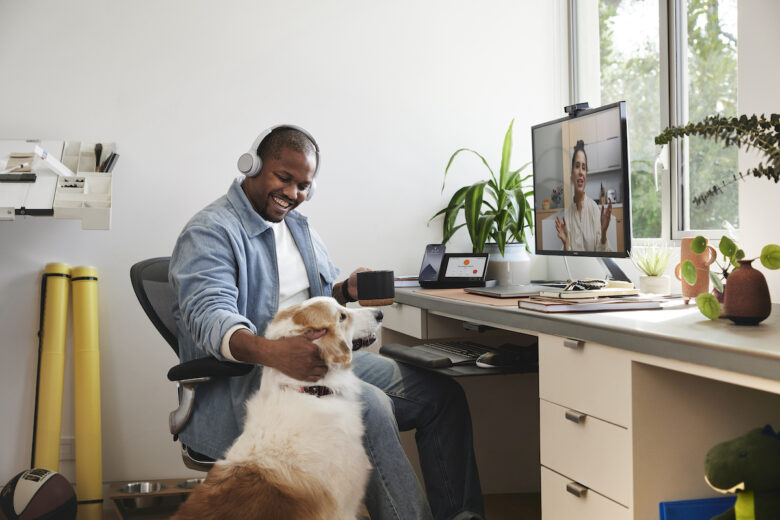 A smiling professional in a denim jacket and a headset takes a moment to pet his dog during a video meeting via Webex