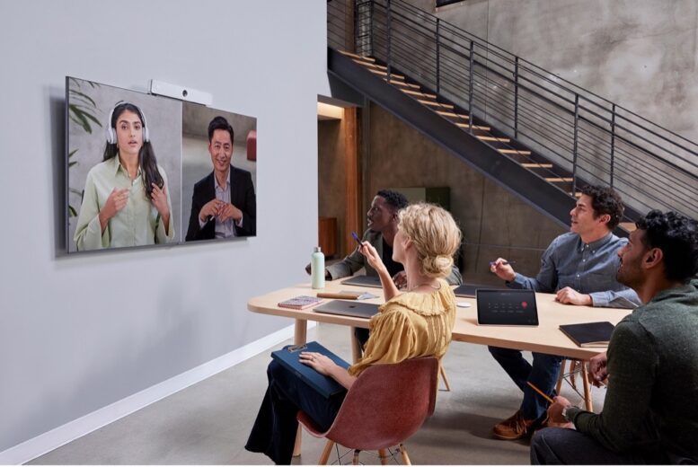 Four people sit at a table, interacting with their colleagues on a video conferencing device mounted on the wall