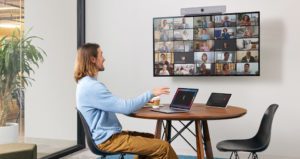 A person sits at a desk, talking to many others who appear via a wall-mounted video conferencing monitor