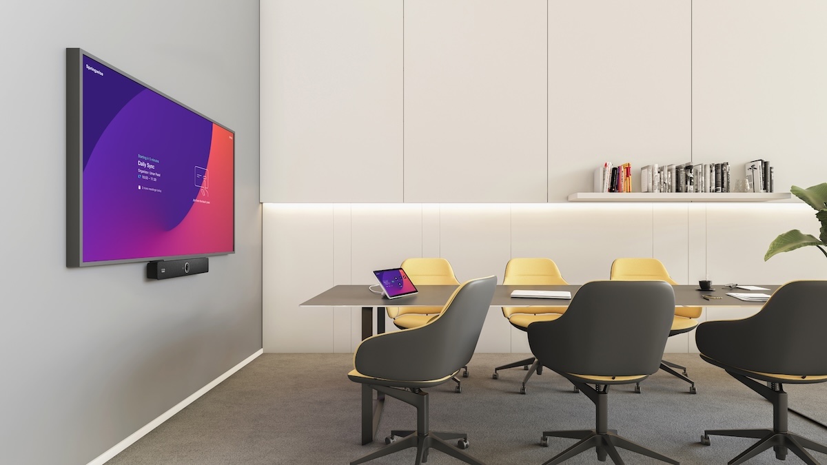 A modern small conference room featuring a large wall-mounted display. A dark grey conference table with several yellow and grey chairs is in the foreground.