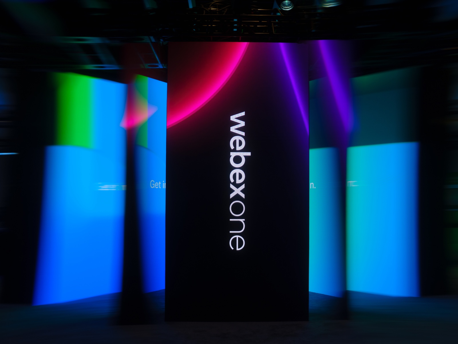 A dark event stage featuring illuminated WebexOne display panels in blue, purple, and pink, with the WebexOne logo prominently displayed on a central vertical screen.