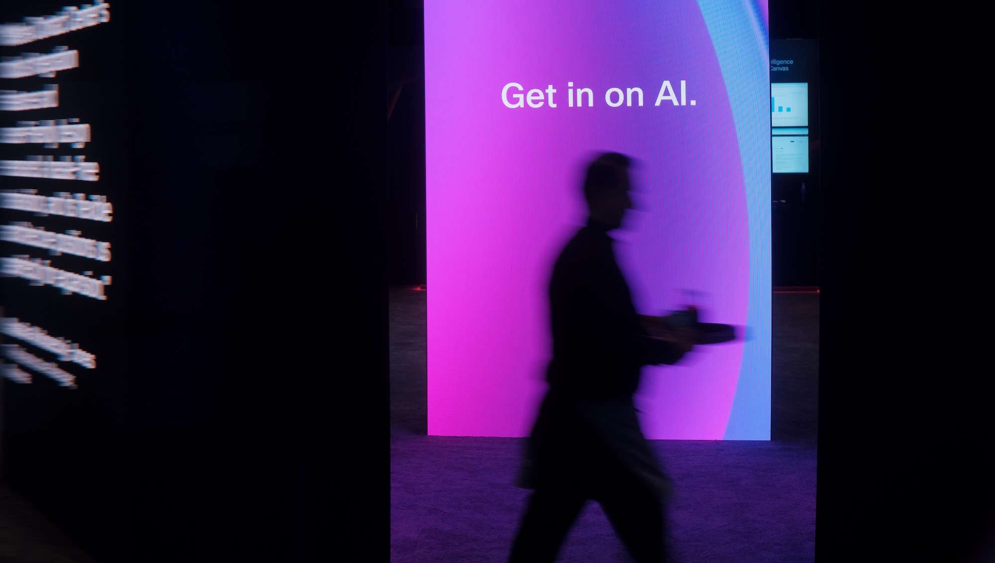 Blurry silhouette of a person carrying a tray walks past a large magenta-and-blue illuminated screen displaying “Get in on AI.” in a dark exhibition space.