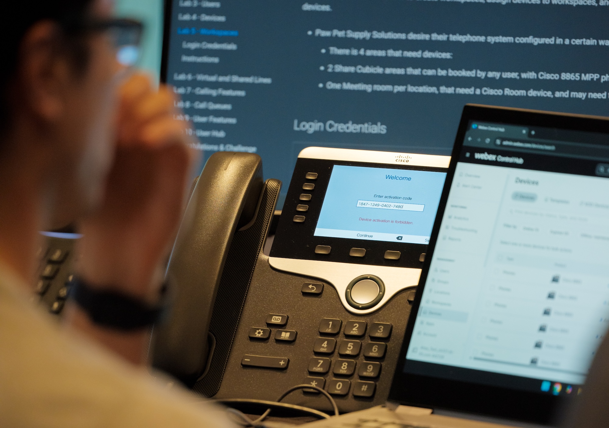 A blurred person sits beside a Cisco desk phone displaying an activation screen, with a laptop showing Webex device settings in the foreground and setup instructions on a monitor behind it.