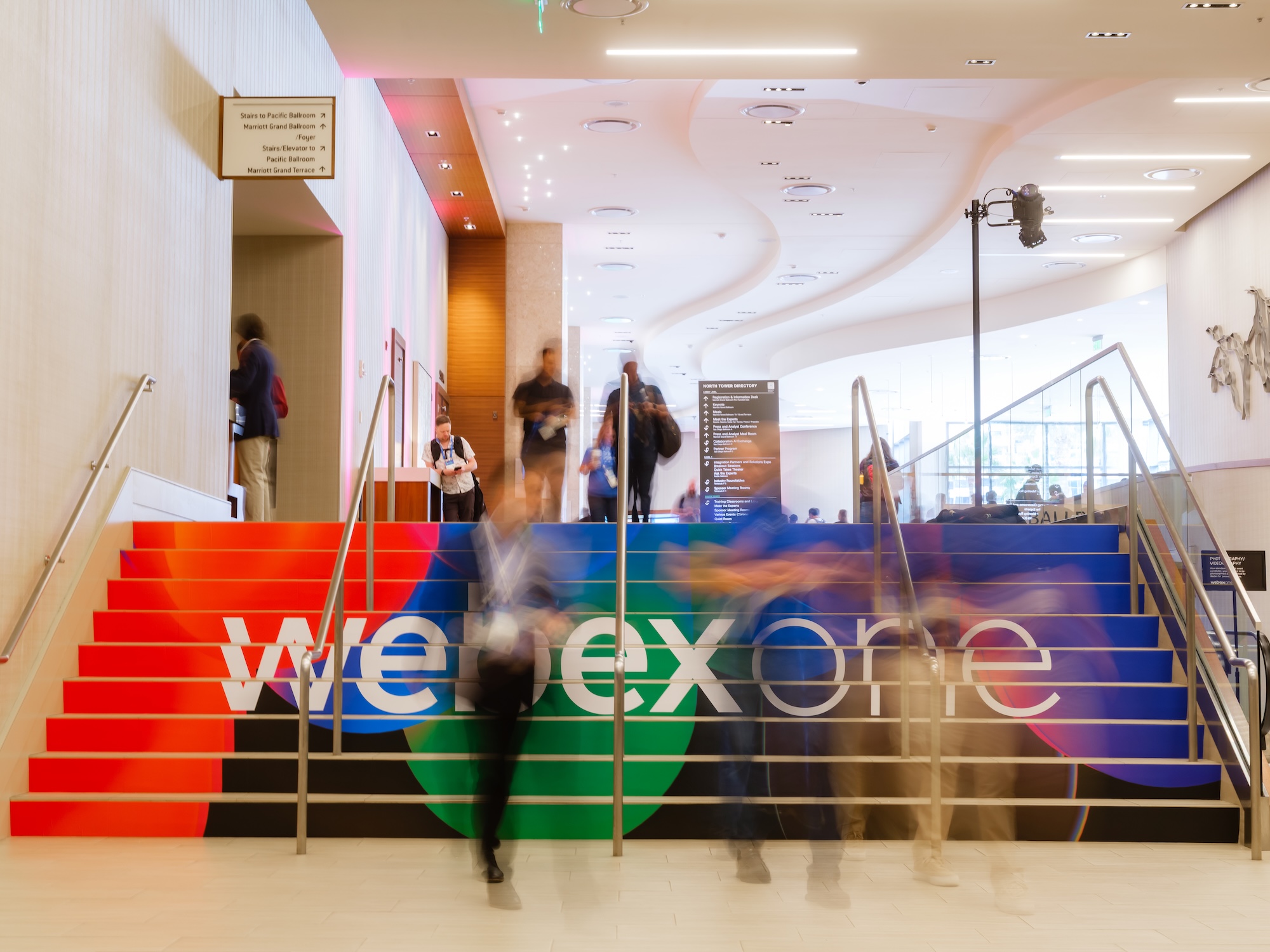 WebexOne attendees move through a conference venue near a colorful staircase featuring the WebexOne logo.